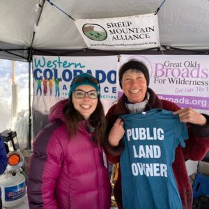 Women in winter jackets, smiling, holding a t-shirt that says "Public Land Owner"